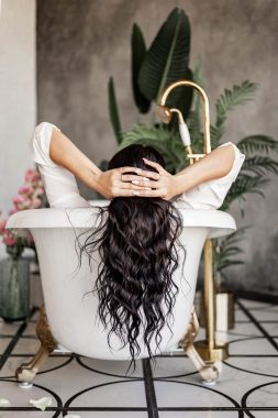Back view fashion curly brunette female lying in bath with water relaxing enjoying spa procedure Back view fashion curly brunette female lying in bath with water relaxing enjoying spa procedure. Glamorous lady in white male shirt resting at luxury residential apartment interior retro bathroom